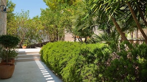 Un chemin bordé de haies verdoyantes et de palmiers, qui mène à un bâtiment à l’architecture en pierre.