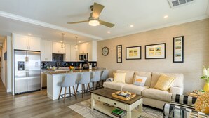 Una cocina moderna con una isla central, un comedor con taburetes de bar, un sofá con cojines decorativos y una mesa de centro.