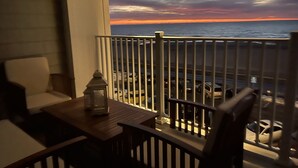Un balcón con vista al atardecer sobre el océano, una mesa de madera, una silla y un farol.