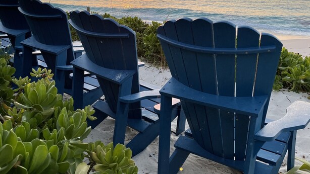 A row of blue Adirondack chairs facing the ocean at sunset.