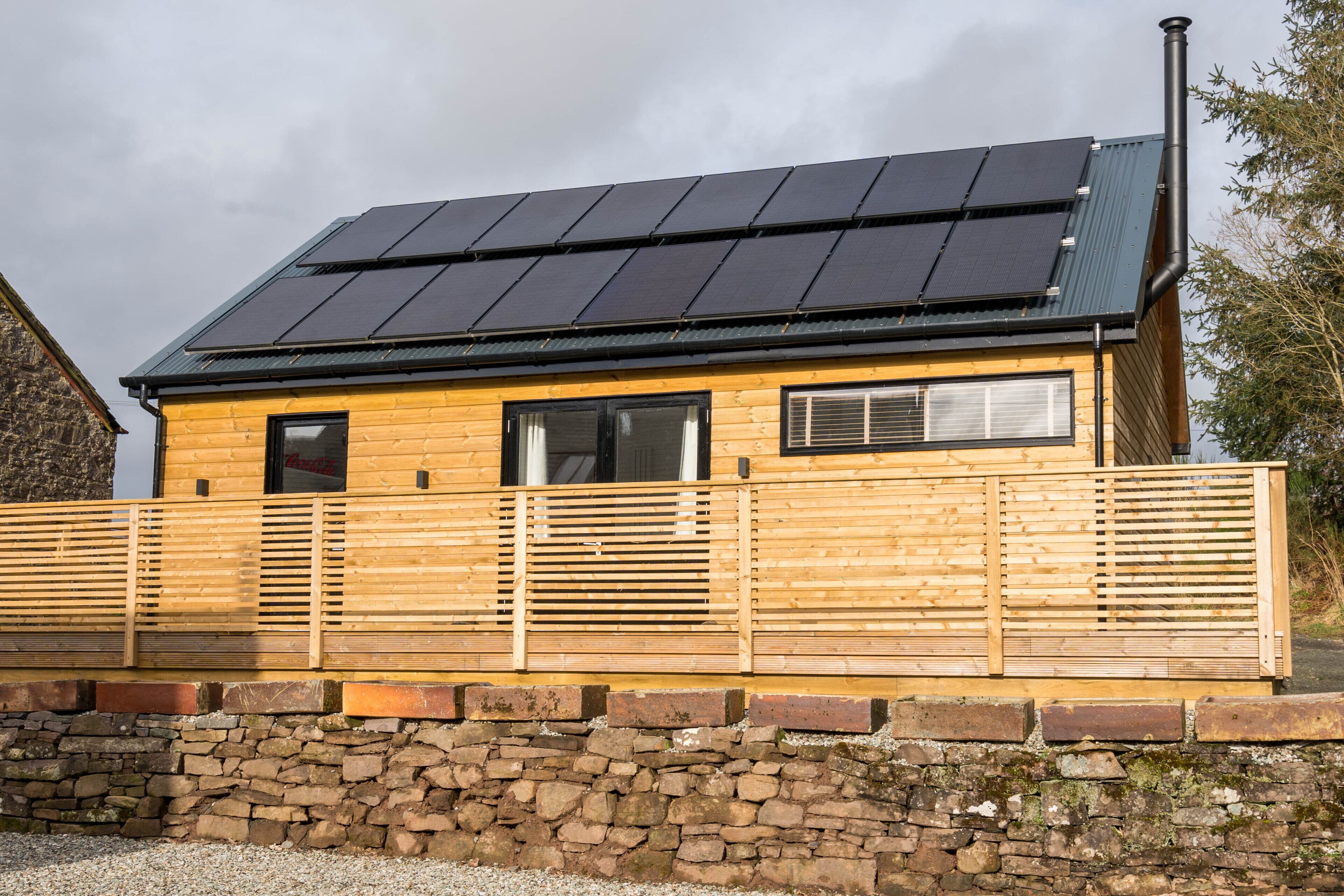 A wooden cabin with a stone foundation, solar panels on the roof, and a stone wall in front.