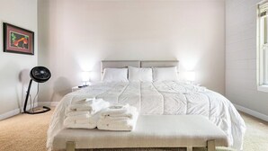 Una cama bien hecha con ropa de cama blanca y un banco al pie, un ventilador de pie y un cuadro en la pared.