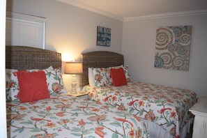 Une chambre à coucher avec une couvre-lit à motifs floraux, deux coussins rouges, une tête de lit en osier, une lampe de chevet et une œuvre d’art murale.