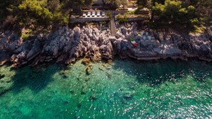 Eine Luftaufnahme einer Küstenimmobilie mit einer Steinstufe, die zu einer Terrasse führt, umgeben von Bäumen und klarem, türkisblauem Wasser.