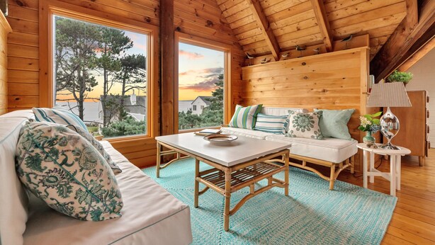 Une pièce chaleureuse avec un plafond en bois, un canapé agrémenté de coussins à motifs, une petite table et une vue sur les arbres et un coucher de soleil, le tout visible à travers de grandes fenêtres.