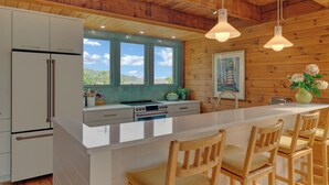 Une cuisine moderne avec des armoires blanches, un grand îlot central doté de tabourets, des poutres en bois au plafond et une vue sur l’extérieur par une fenêtre.