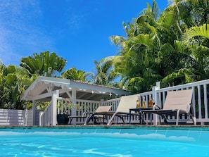 A poolside area with lounge chairs, a covered seating area, and lush tropical foliage.