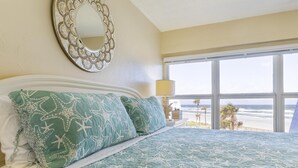 Quarto com cama grande, espelho decorativo e vista para a praia através de uma janela ampla.