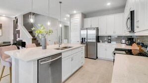 Une cuisine moderne dotée d’un îlot central, d’appareils électroménagers en acier inoxydable, de luminaires suspendus et d’un escalier menant à un autre niveau.