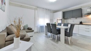 A modern living room with a sofa, a dining table with chairs, a kitchen area with cabinets and a TV, and a window with curtains. - Holiday Home Vesna with pool and children area (Suhovare)