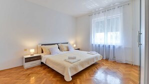 A bedroom with a wooden herringbone floor, a bed with white linens, a nightstand with a lamp, and sheer curtains on the window. - Holiday Home Vesna with pool and children area (Suhovare)