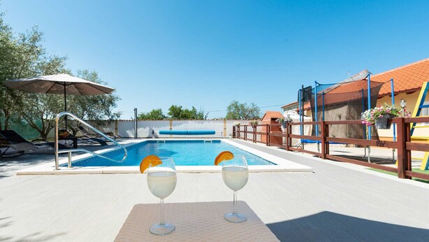 A poolside area with a clear blue pool, two glasses of a beverage with orange slices, and a wooden fence. - Holiday Home Vesna with pool and children area (Suhovare)