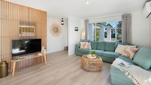 Ein modernes Wohnzimmer mit Fernseher, einem grünen Sofa, einem runden Kaffeetisch aus Korbmöbeln und einer Holzabtrennung.
