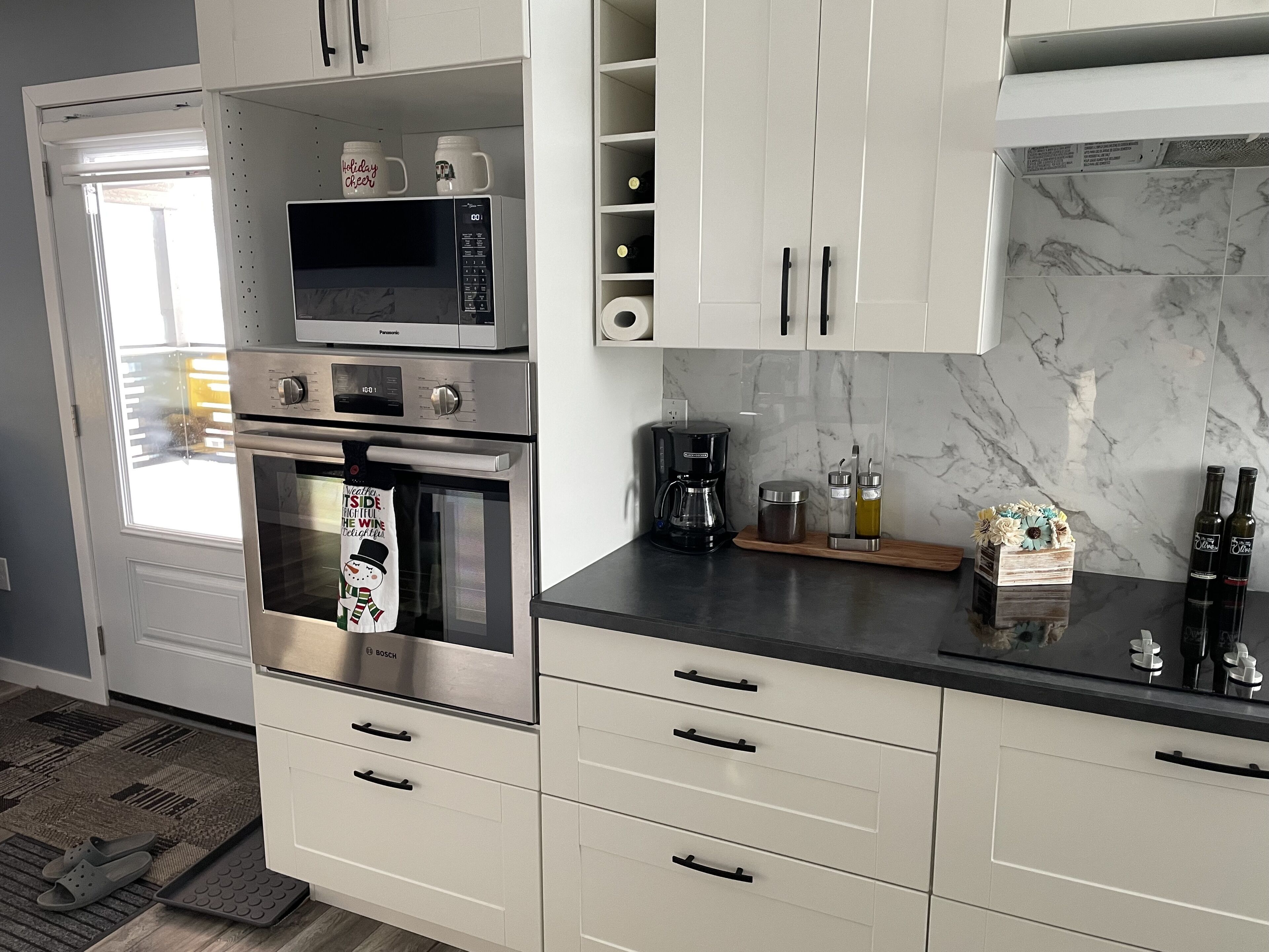 Eine Küche mit integriertem Mikrowellenherd, Backofen und Kaffeemaschine. Es gibt Schränke mit Griffen, eine Arbeitsplatte mit Kaffeemaschine und eine dekorative Schachtel mit Blumen.