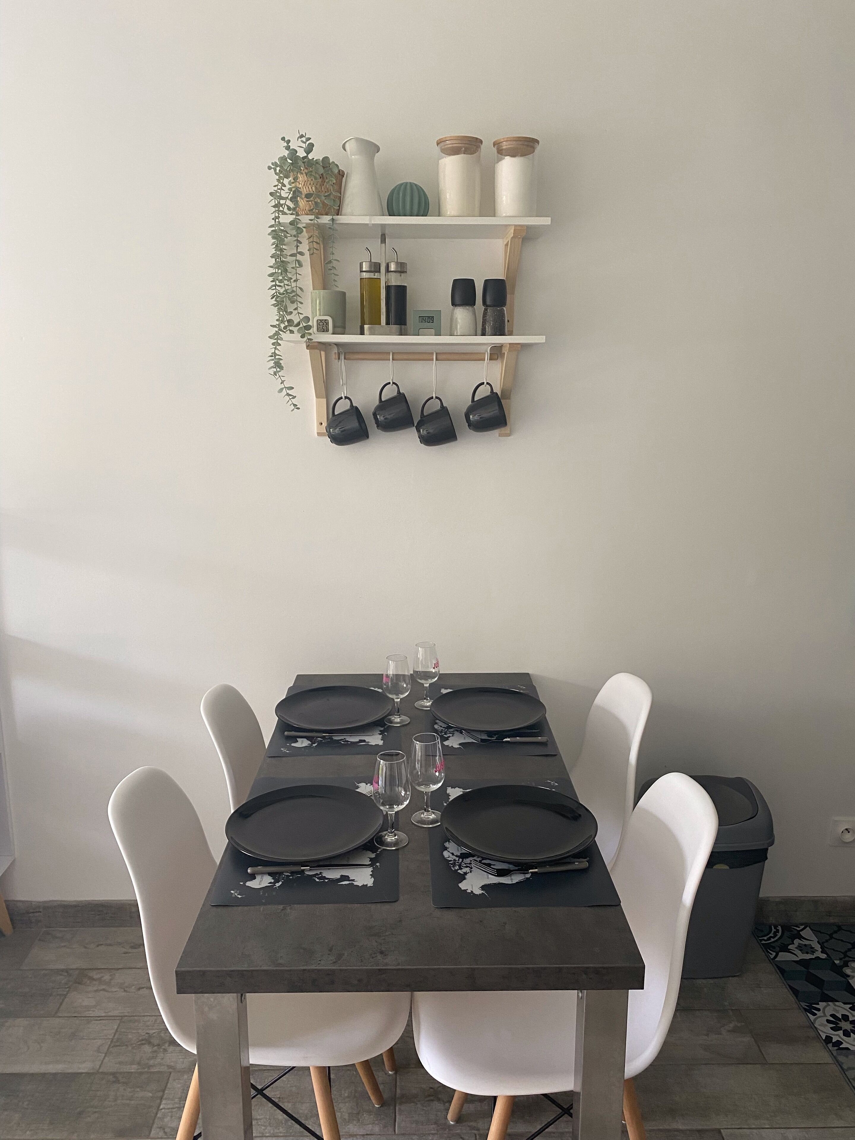 Mesa de jantar posta com pratos pretos e copos de vinho, cercada por cadeiras brancas. Acima da mesa, há uma estante com potes, uma planta e sacos pretos pendurados.
