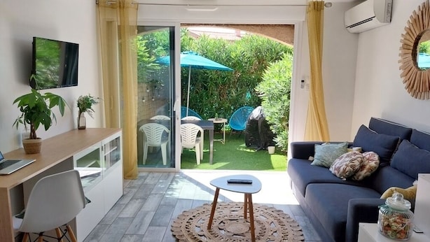 A modern living room with a sofa, a coffee table, and a desk with a laptop. There is a sliding glass door leading to a patio with outdoor furniture and greenery. - Studio 100m from the beach, patio, car park, air conditioning (Six-Fours-les-Plages)