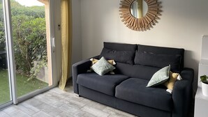 A modern living room with a dark-colored sofa, a round mirror with a wooden frame, and a sliding glass door leading to a balcony with greenery. - Studio 100m from the beach, patio, car park, air conditioning (Six-Fours-les-Plages)