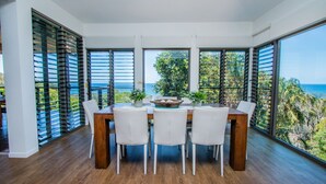 Ein Essbereich mit Holztisch, weißen Stühlen, großen Fenstern mit Jalousien und Blick auf das Meer.