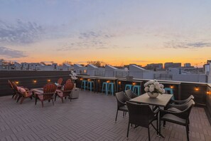 Une terrasse sur le toit avec des sièges extérieurs, un espace bar et une vue sur les toits de la ville au coucher du soleil.