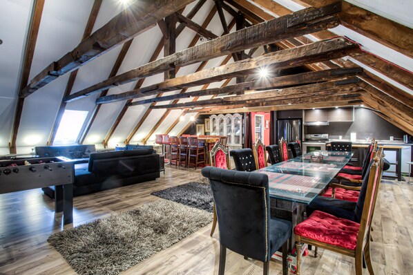 A spacious loft with exposed wooden beams, a dining area with a glass table, and a cozy seating area with a foosball table.