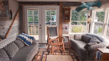 Relax in a cozy room with a large window offering a view of the outdoors, a comfortable sofa, a wooden rocking chair, and a bookshelf filled with books.