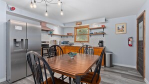 Cocina moderna con refrigerador de acero inoxidable, gabinetes de madera, mesa de comedor con sillas y espacio para café.