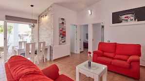 Un salon avec un canapé rouge, des fauteuils blancs, une table basse en verre et un mur en briques.