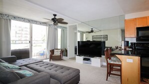 A modern living room with a large grey sectional sofa, a flat-screen TV, and a dining area with wooden cabinets and a black microwave. - Spacious 2br Waikiki (Honolulu)
