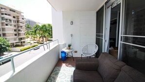 A balcony with a sofa, a small table, and a chair. There is a view of a street and buildings in the background. - Spacious 2br Waikiki (Honolulu)