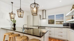 Une cuisine moderne dotée d’un îlot central, de tabourets de bar, de luminaires suspendus et offrant une vue sur l’extérieur grâce à une fenêtre munie de stores.