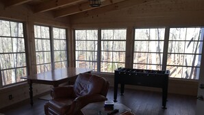 A cozy room with a leather sofa, a wooden table, and a foosball table, with large windows offering a view of trees.