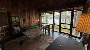 A rustic cabin interior with a wooden dining table, benches, and a view of a forest through large windows.