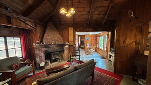 A rustic cabin interior with a fireplace, wooden walls, a dining area, and a view through a window.