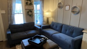 A living room with two dark sofas, a coffee table, and a rug. There is a window with curtains, a lamp, and wall decorations.