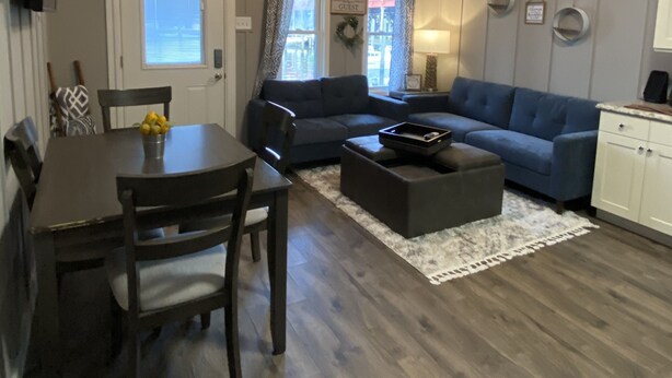 A living room with a dark brown dining table, two dark brown chairs, a dark brown sofa, a dark brown ottoman, and a white rug. There is a ceiling fan, a window with curtains, and a door with a sign that reads "GUEST".