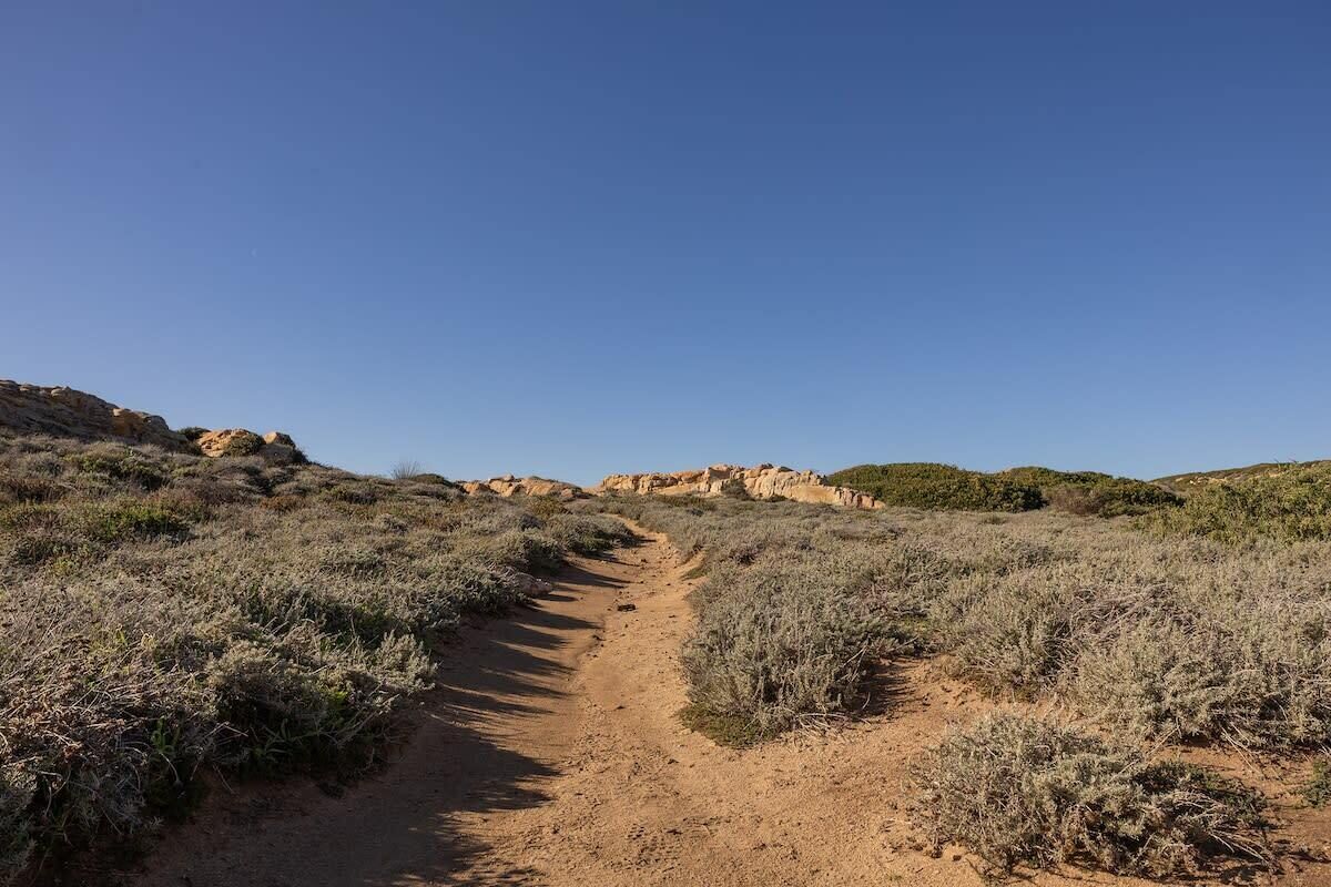 A dirt path leading through a dry, hilly landscape with sparse vegetation.