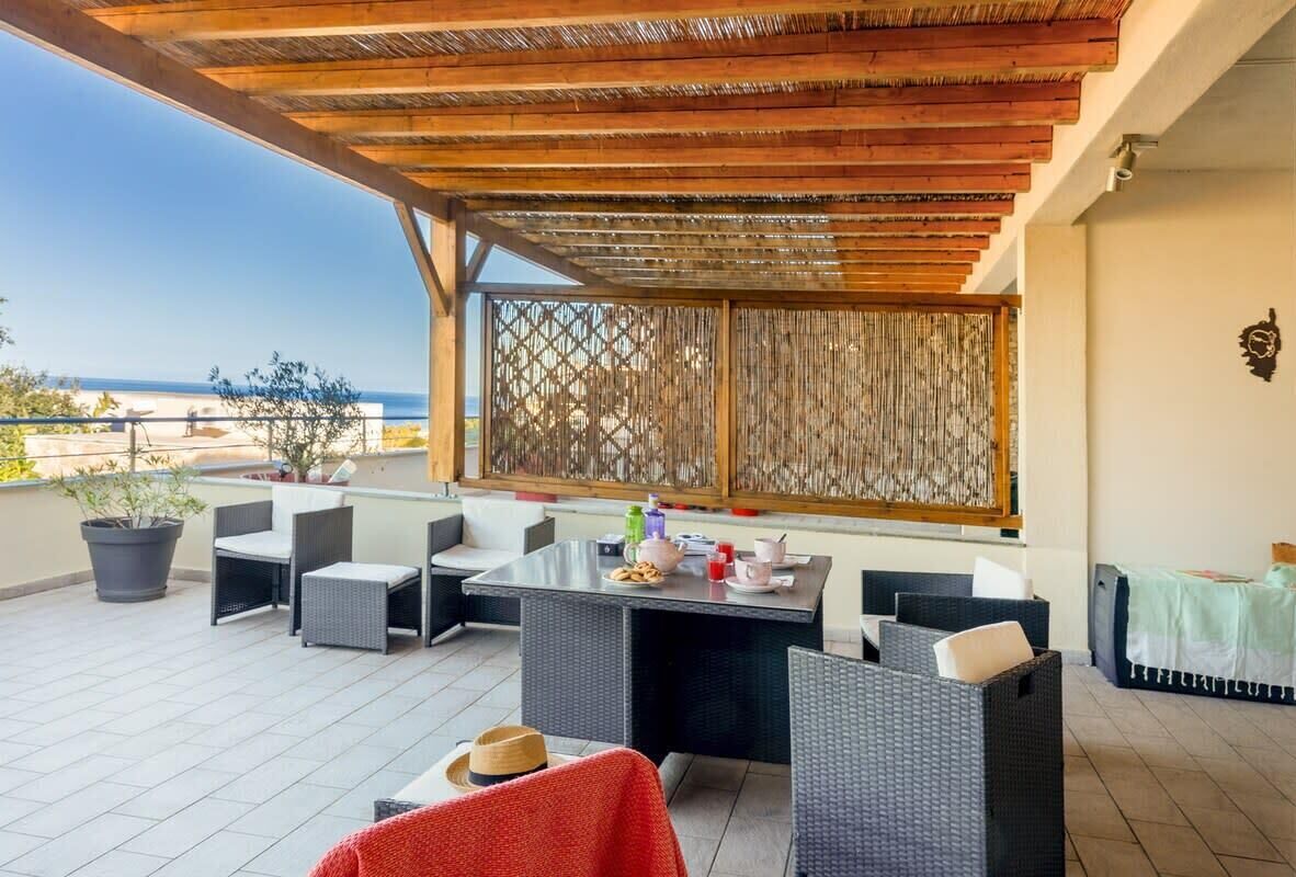 A covered outdoor seating area with wicker furniture, a table with drinks, and a view of the sea.