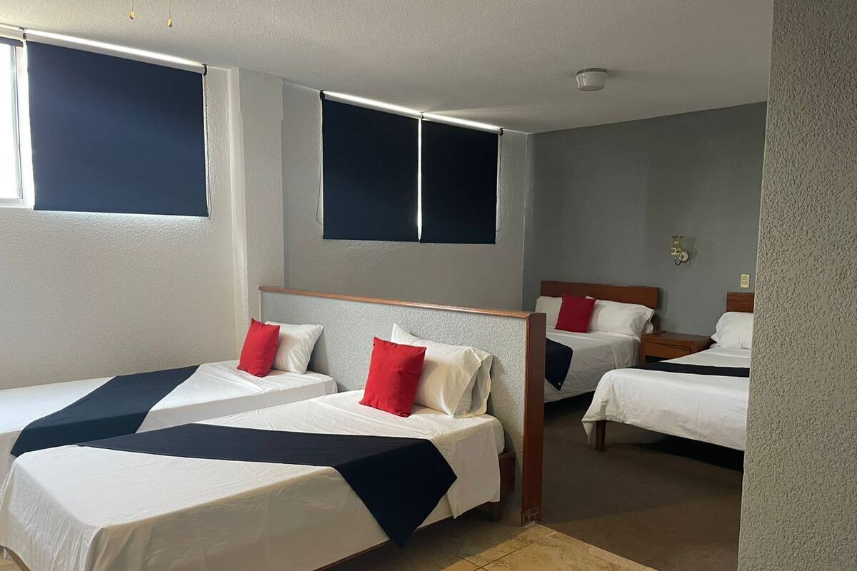 A hotel room with two beds, each with white bedding and red pillows, and a large window with a dark blind.