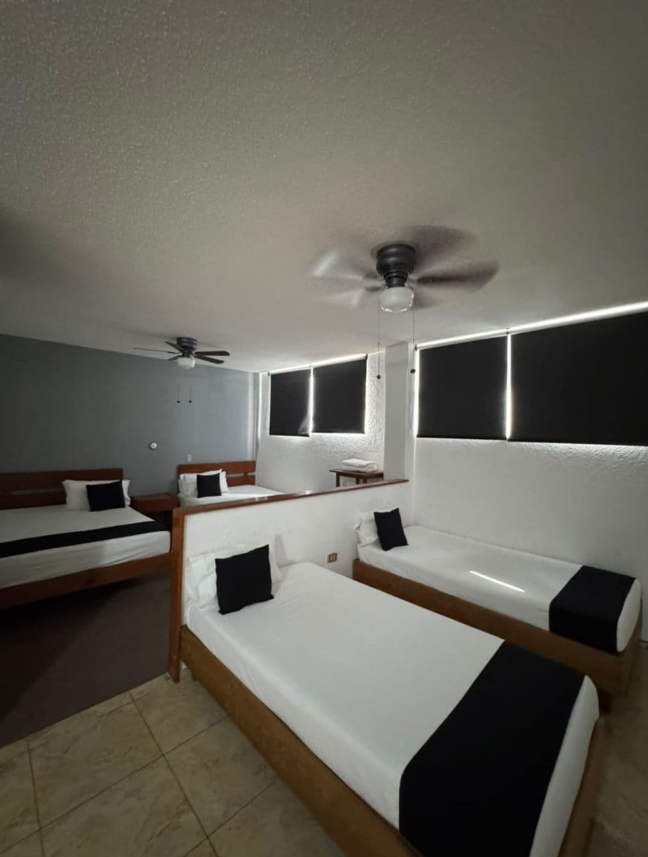 Une chambre d’hôtel avec un grand lit, un lit plus petit, un ventilateur de plafond et une fenêtre avec des stores.