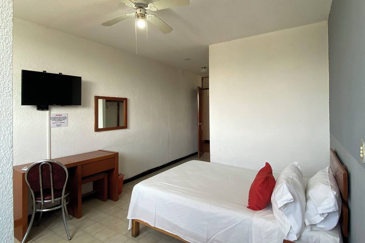 A hotel room with a bed, a desk, a chair, a ceiling fan, a TV, and an air conditioning unit.