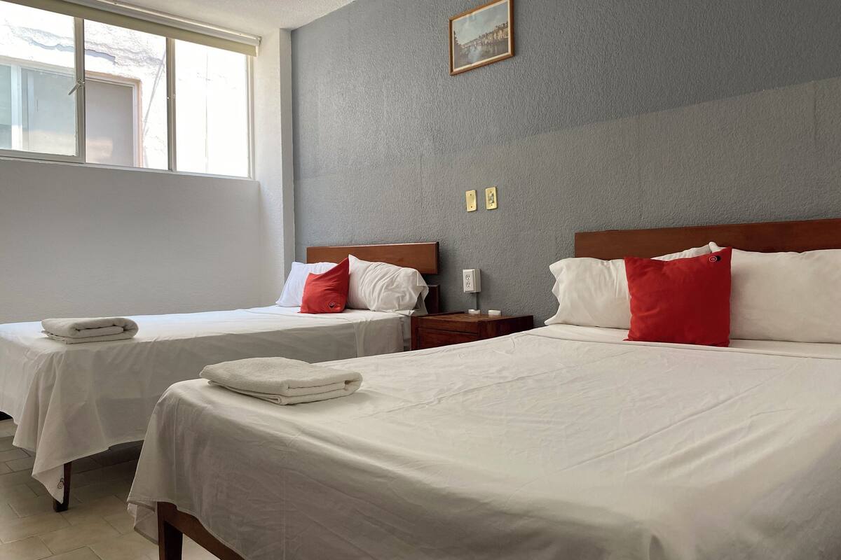 A hotel room with two beds, each with white bedding and red pillows. There is a framed picture on the wall, and a window providing natural light.