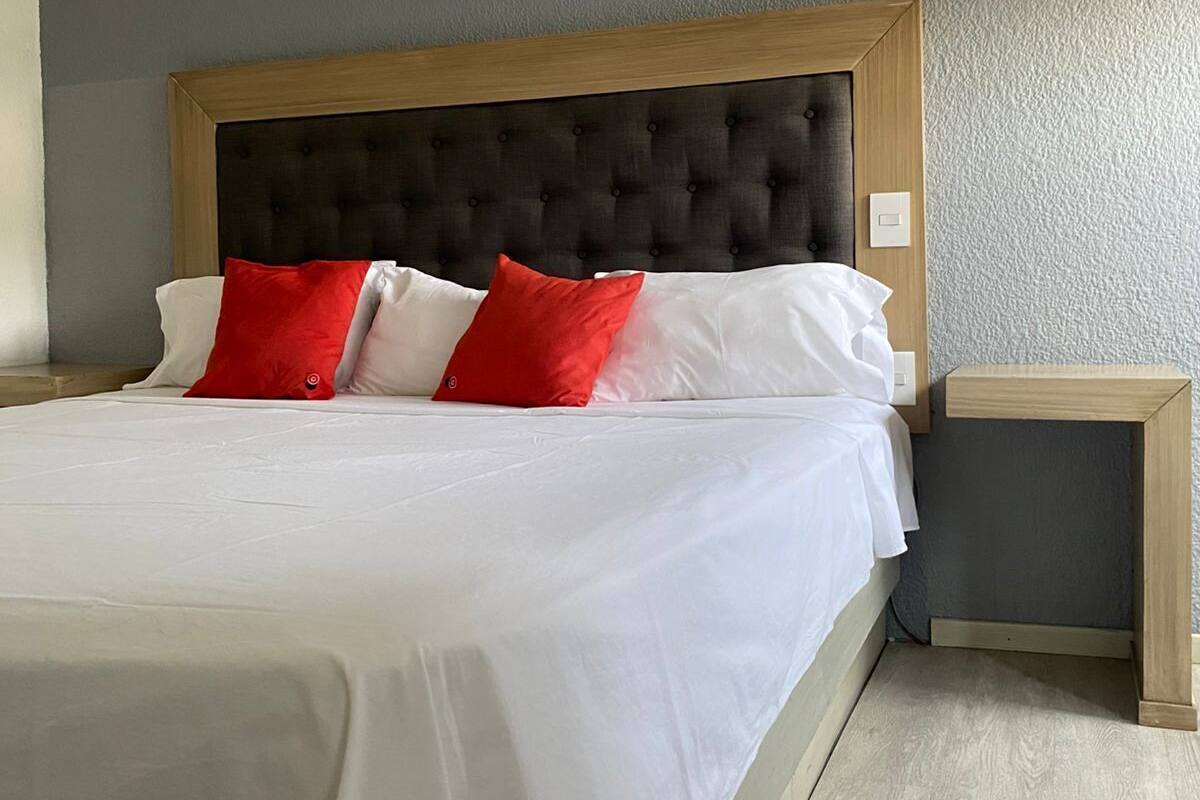 A neatly made bed with white bedding and red pillows, a wooden headboard, and a small bedside table with a light switch.