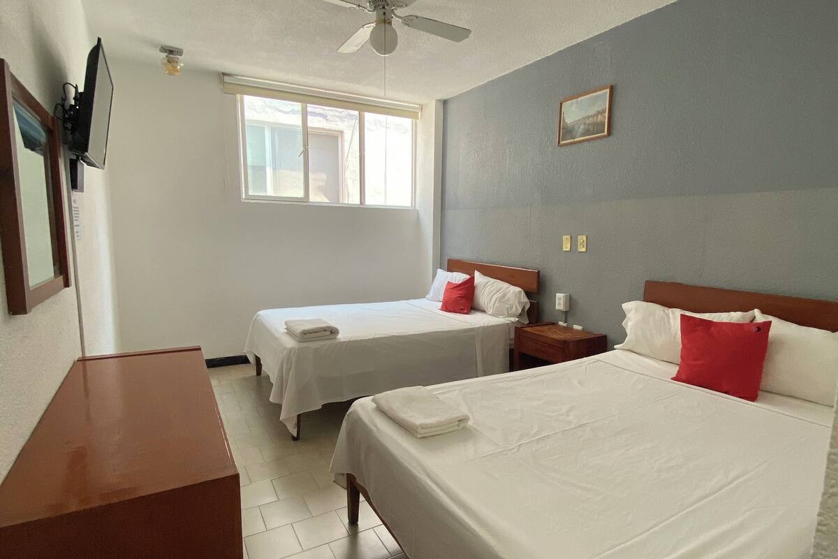 A hotel room with a bed, a desk, a chair, a nightstand, a bedside table, a window, and a ceiling fan.
