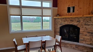 A dining area with a stone fireplace, a large window with blinds, and a view of a grassy field. - Connors Lake Landing - Home/Hangar & Federal License Grass Runway - 2WS2 (Webster)