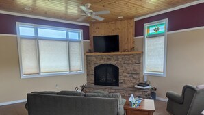 A living room with a stone fireplace, a flat-screen TV mounted above it, and a ceiling fan. - Connors Lake Landing - Home/Hangar & Federal License Grass Runway - 2WS2 (Webster)