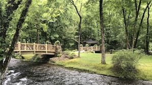 A wooden bridge over a flowing river, surrounded by lush greenery and a stone building in the background.