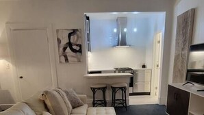 Un salon moderne avec un canapé, un pouf et un téléviseur posé sur un meuble. On aperçoit une cuisine au fond.