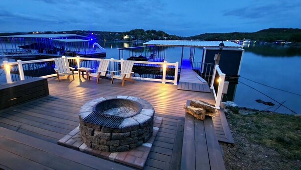 Eine Holzterrasse mit einem Steinkamin, Sitzgelegenheiten im Freien und Blick auf einen Yachthafen im Abendrot.
