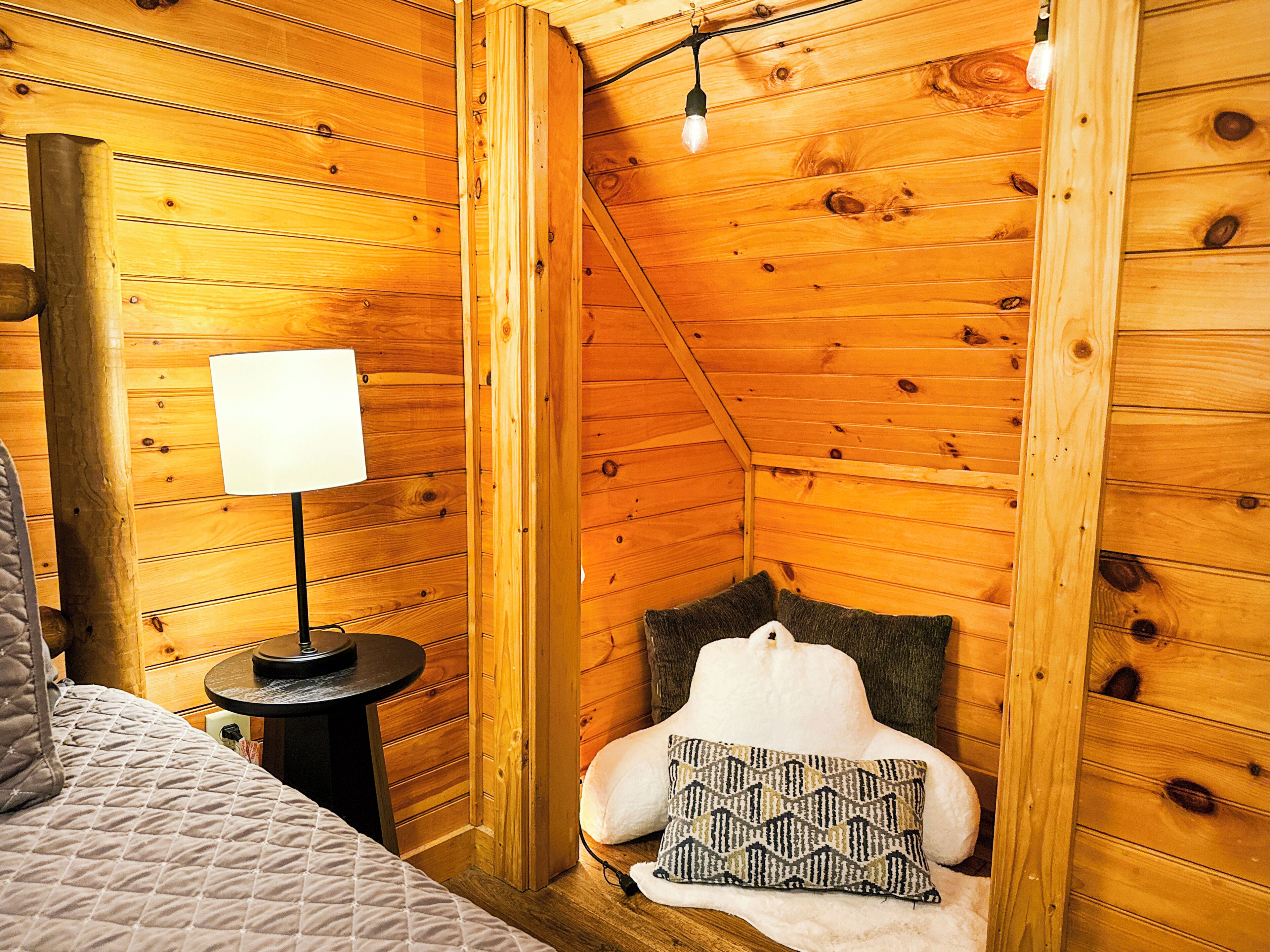 Une pièce chaleureuse avec des murs en bois, un lit, une petite table avec une lampe et un coussin sur une chaise.