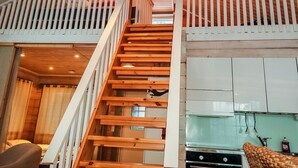 Una escalera de madera lleva a una zona de dormir en un altillo, con una cocina al fondo.